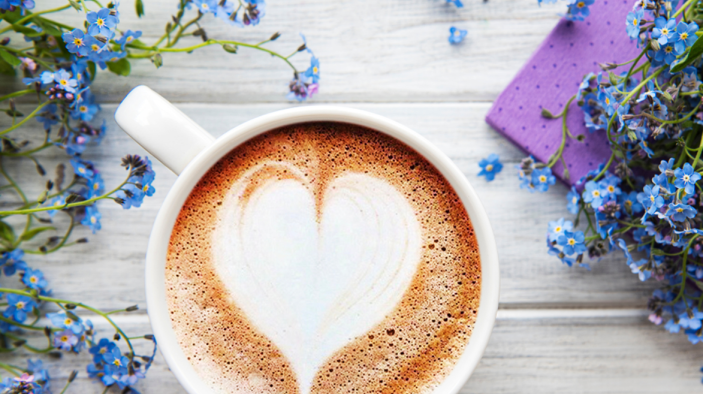 Overhead view of a coffee cup with a heart made out of milk foam sat on a table surrounded by forget me not flowers