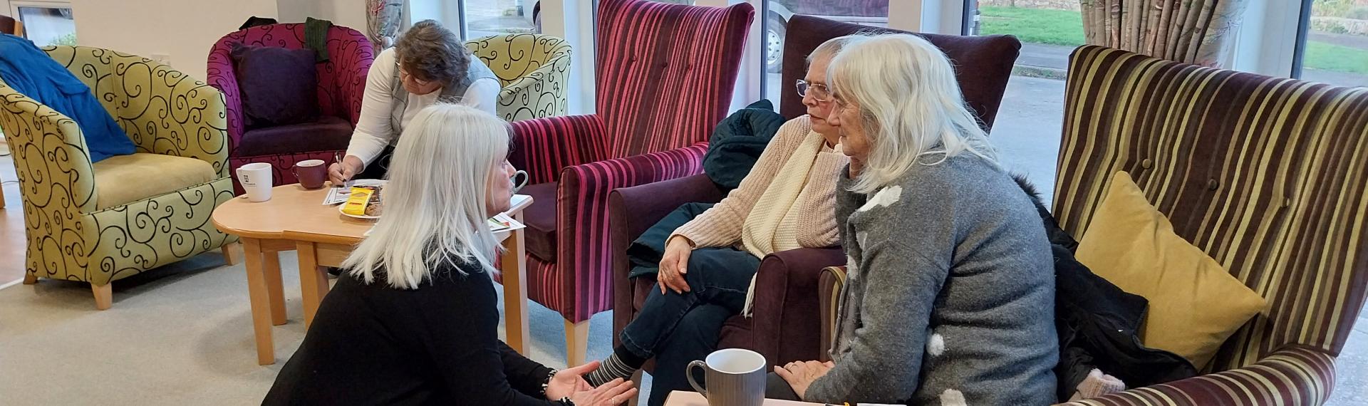 Three ladies sat on colourful armchairs with another lady crouched down on the floor talking to them